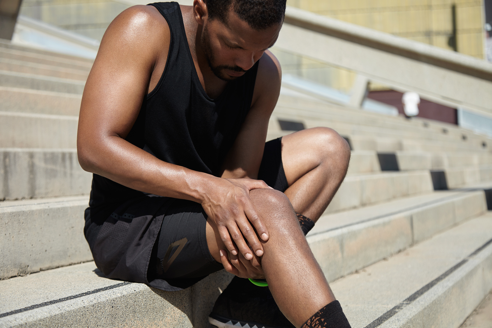 Handsome muscular male jogger wearing black training outfit touching his knee in pain with clasped hands having sprain or rupture in his muscles after exercising outdoors. Sports injury concept
